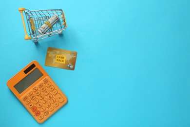 Photo of Calculator, credit card and rolled dollar banknotes in shopping cart on light blue background, flat lay with space for text. Cashback concept