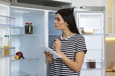 Young woman writing notes near empty refrigerator in kitchen