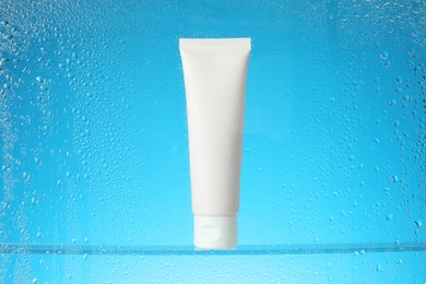 Photo of Tube with moisturizing cream on light blue background, view through wet glass