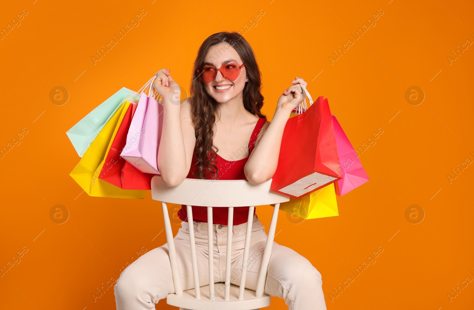 Photo of Happy woman in stylish sunglasses with many colorful shopping bags on chair against orange background