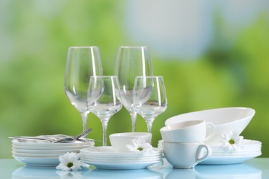 Set of many clean dishware, cutlery, flowers and glasses on light blue table against blurred green background