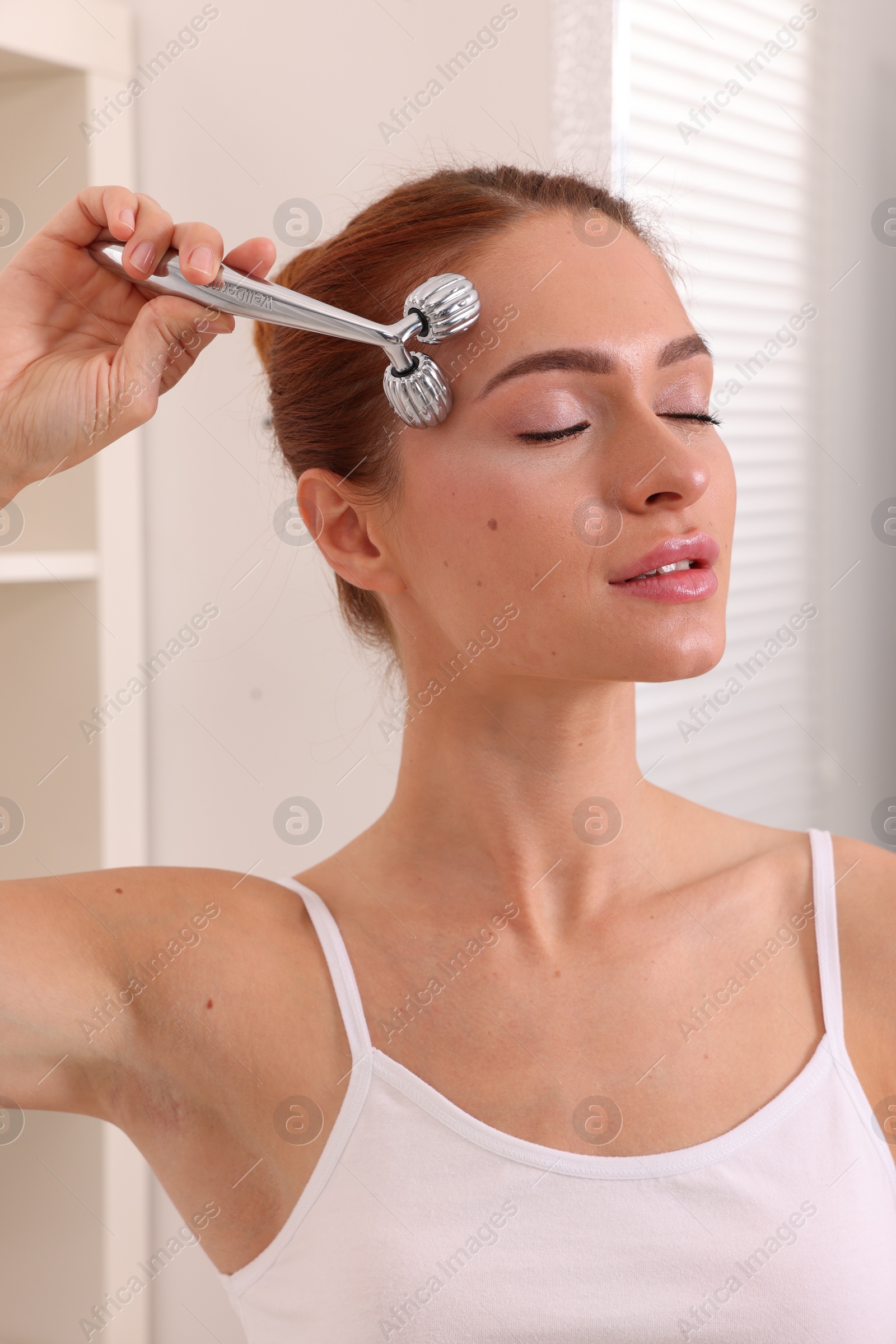 Photo of Young woman massaging her face with metal roller in bathroom