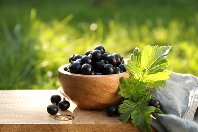 Ripe blackcurrants in bowl and leaves on wooden surface outdoors. Space for text