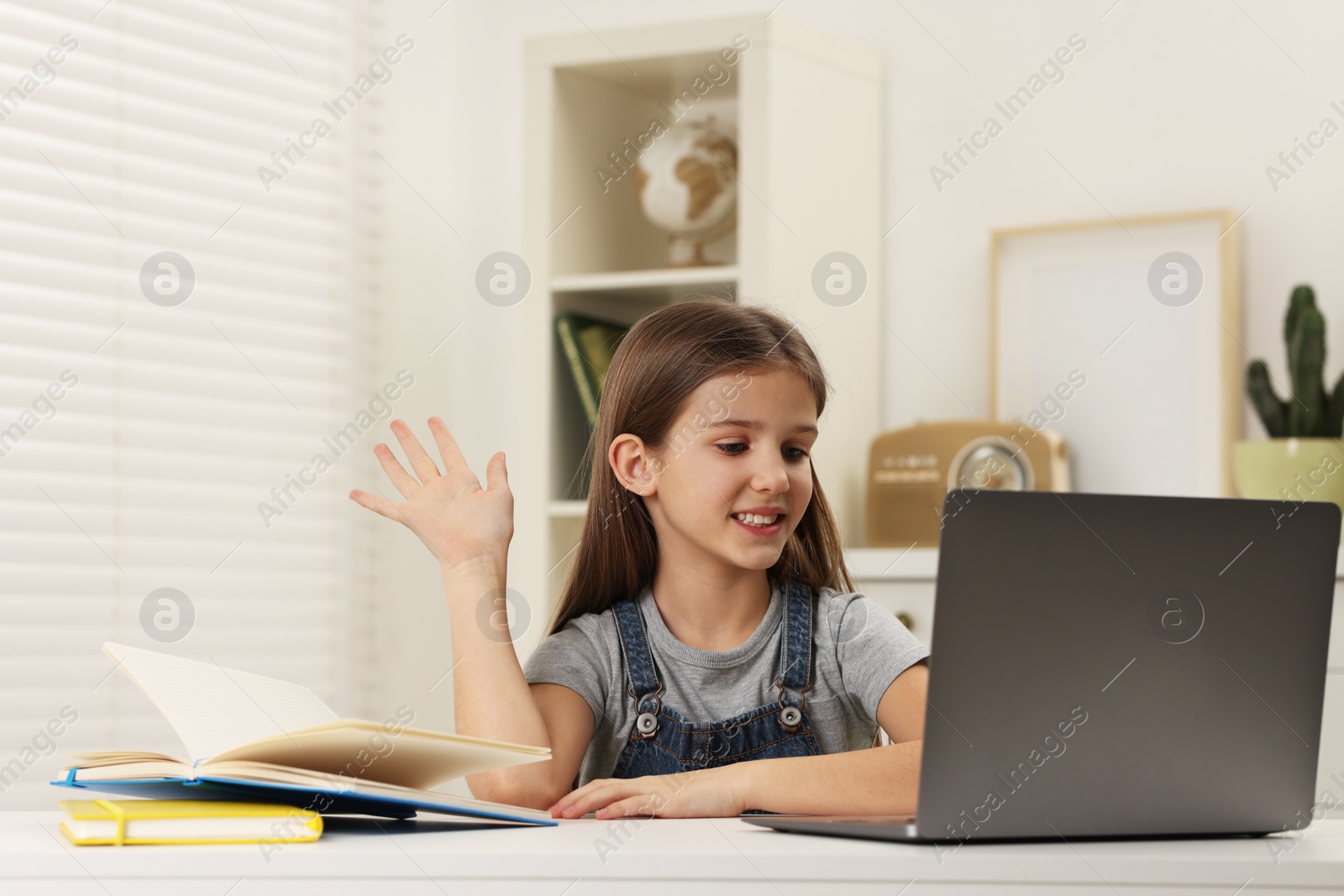 Photo of Cute girl waving hello during online lesson via laptop at white table indoors