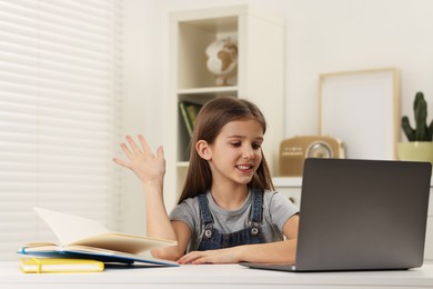 Cute girl waving hello during online lesson via laptop at white table indoors