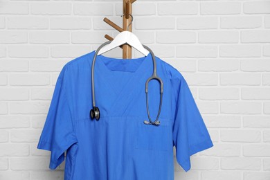 Blue medical uniform and stethoscope hanging on rack near white brick wall