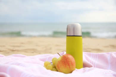 Photo of Metallic thermos with hot drink, fruits and plaid on sandy beach near sea