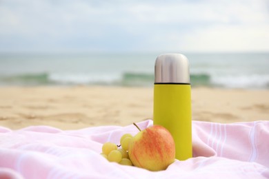 Metallic thermos with hot drink, fruits and plaid on sandy beach near sea