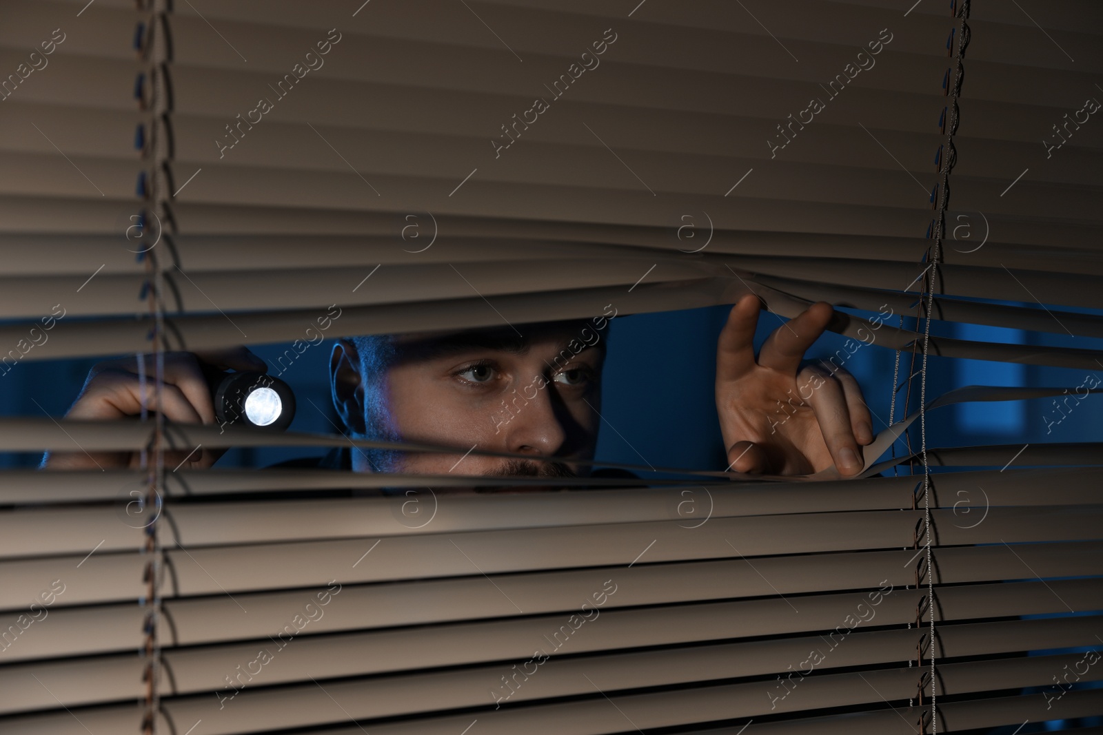 Photo of Male security guard with flashlight looking through window blinds in darkness