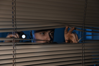 Male security guard with flashlight looking through window blinds in darkness