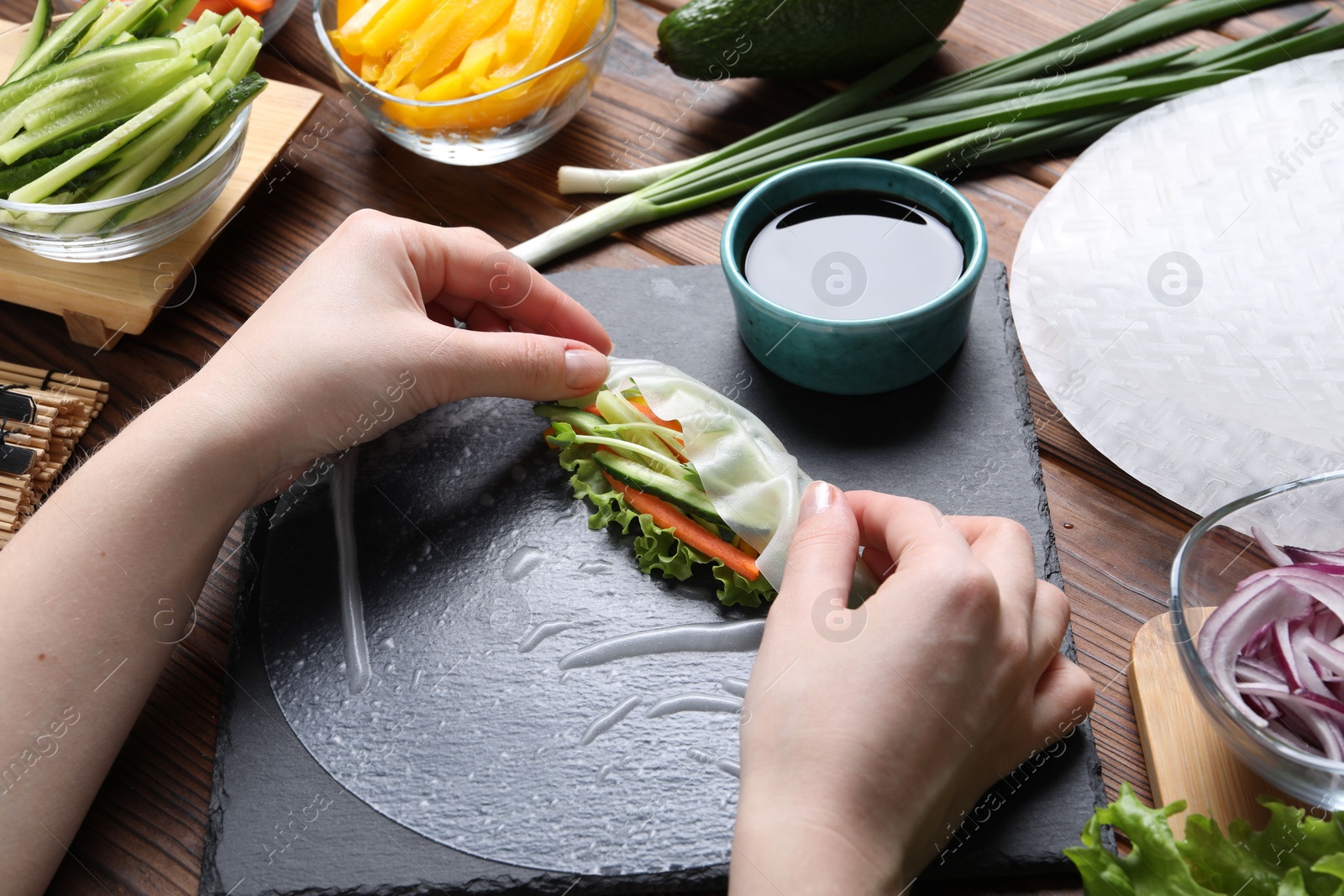 Photo of Woman wrapping spring roll at wooden table with products, closeup