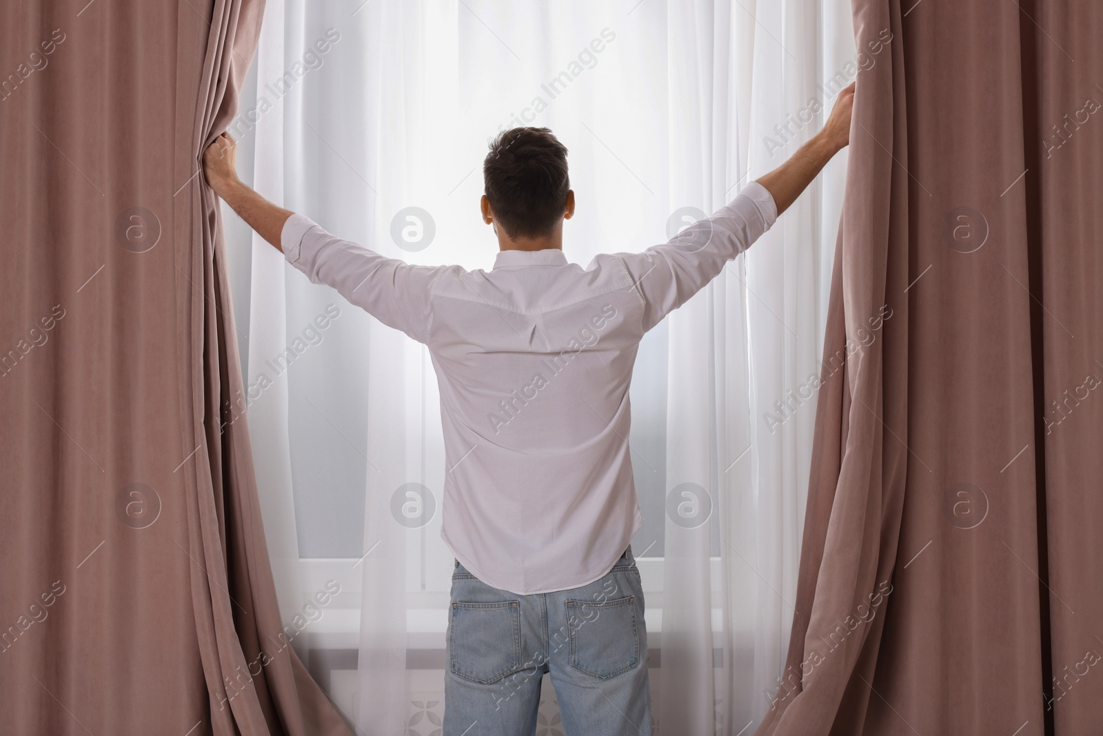 Photo of Man opening window curtains at home, back view