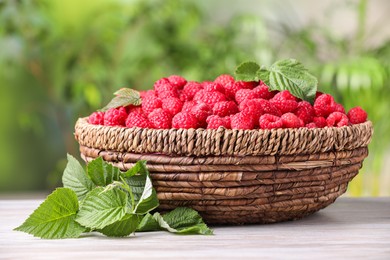 Photo of Wicker basket with tasty ripe raspberries and leaves on white wooden table against blurred green background, closeup