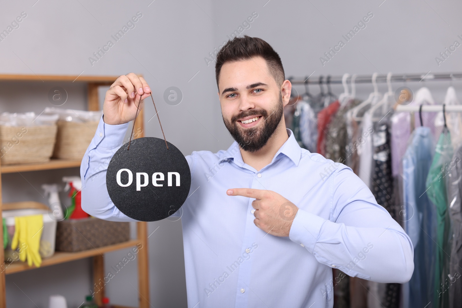 Photo of Dry-cleaning service. Happy worker pointing at Open sign indoors