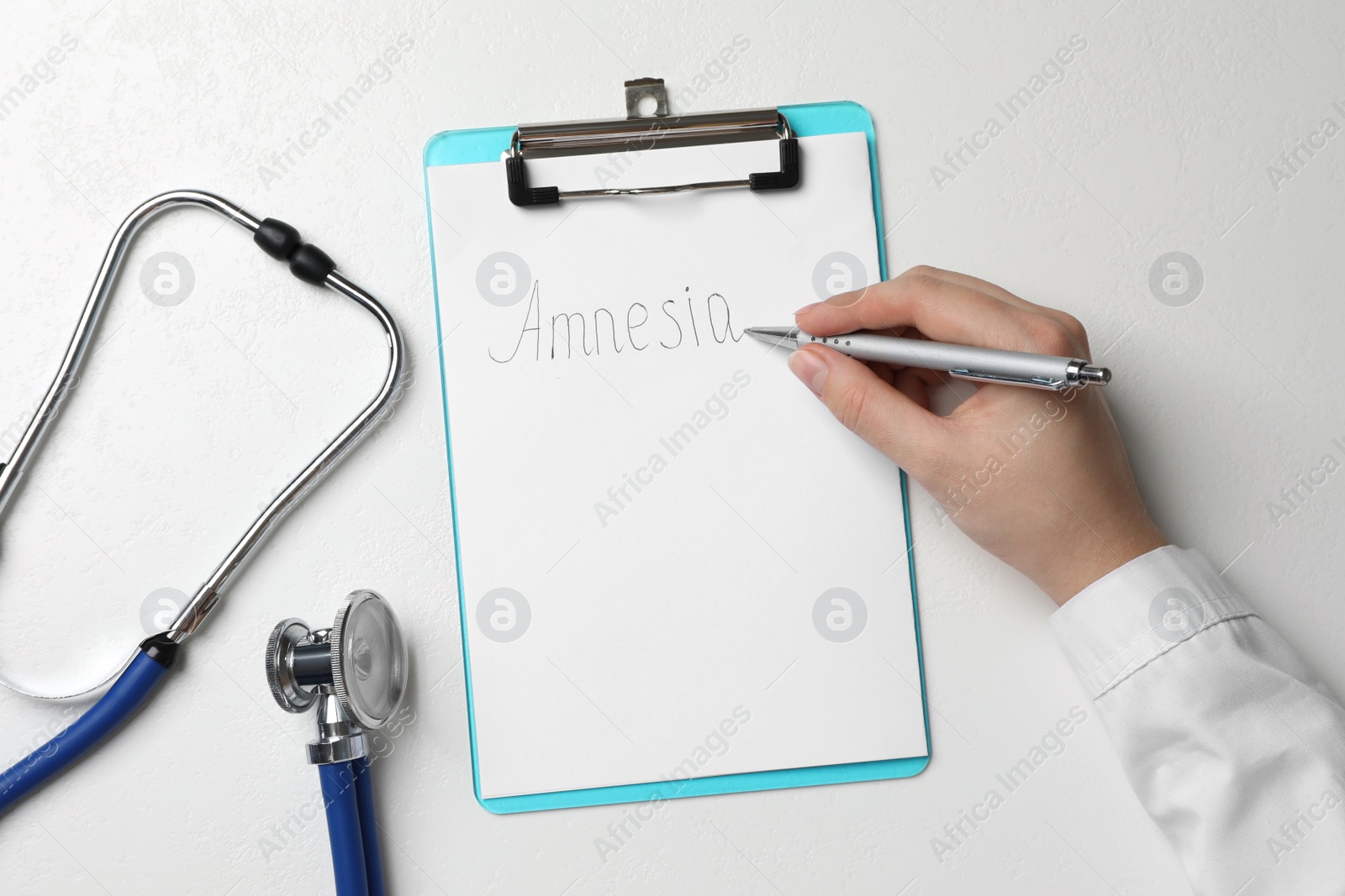 Photo of Woman writing word Amnesia on clipboard at white table, top view