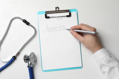 Woman writing word Amnesia on clipboard at white table, top view