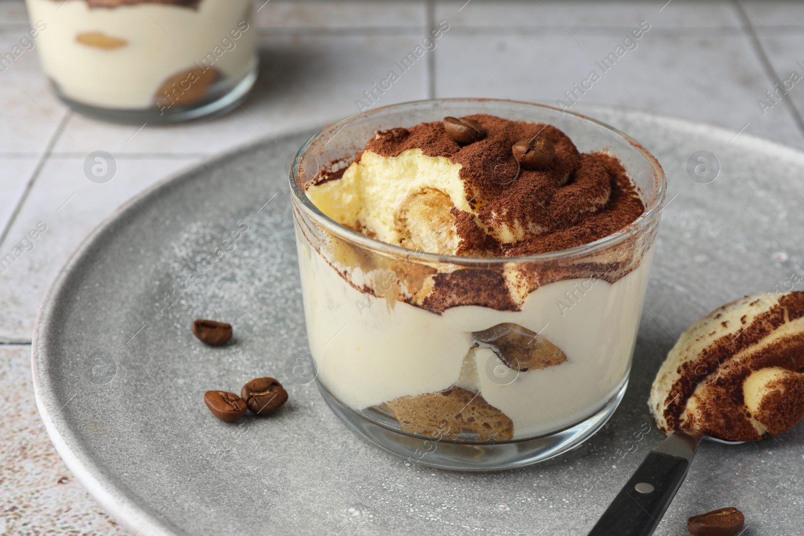 Photo of Delicious tiramisu in glass, coffee beans and spoon on table, closeup