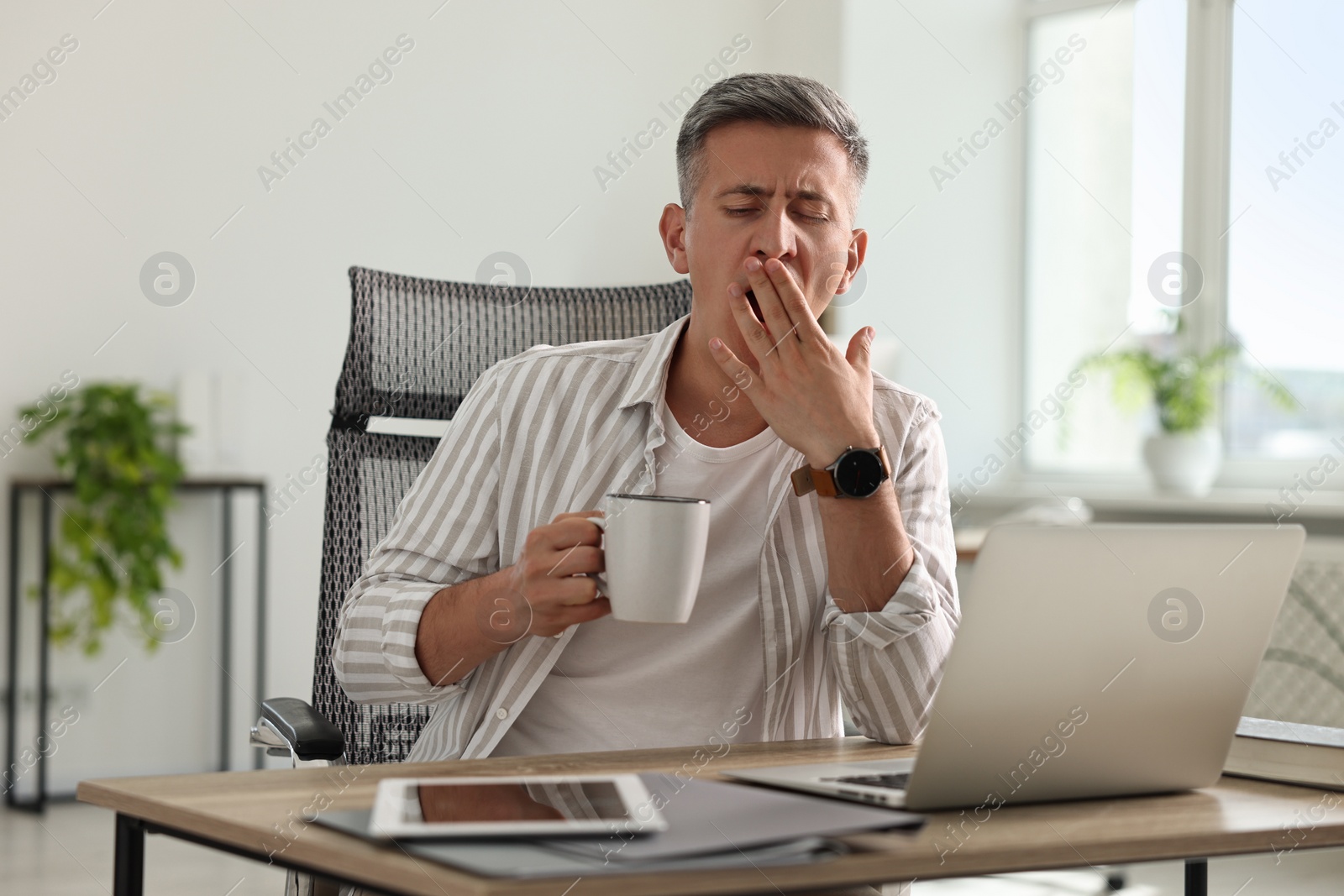 Photo of Man with cup of drink yawning at wooden table in office