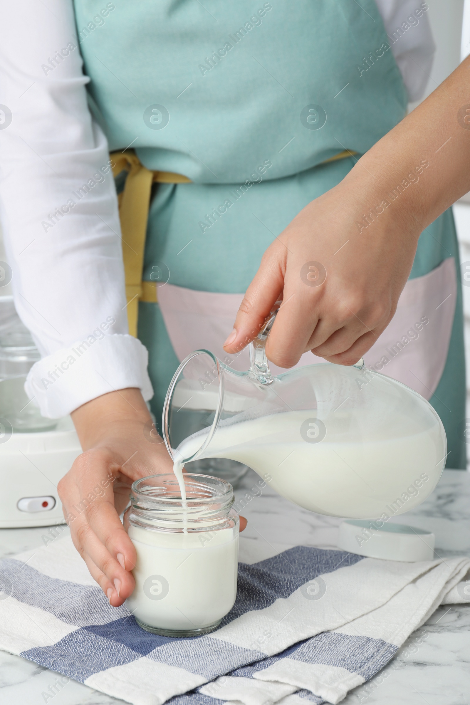 Photo of Woman pouring milk into glass jar at white marble table, closeup. Making yogurt