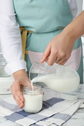 Woman pouring milk into glass jar at white marble table, closeup. Making yogurt