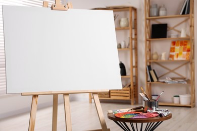 Photo of Wooden easel with blank canvas and different art supplies in studio, closeup