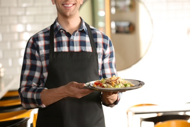 Young waiter in uniform holding tray with tasty dish at workplace