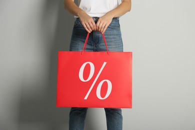 Woman holding paper shopping bag with percent sign on light grey background, closeup. Discount concept