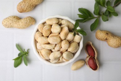 Fresh peeled peanuts in bowl and leaves on white tiled table, flat lay