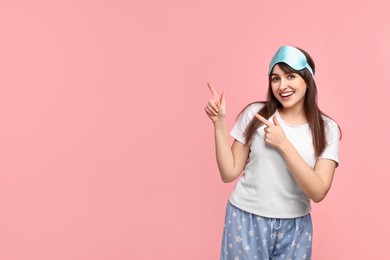 Photo of Happy woman in pyjama and sleep mask pointing at something on pink background, space for text