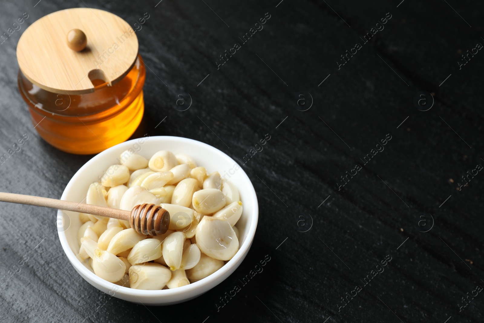 Photo of Peeled garlic cloves in bowl and honey on black table. Space for text