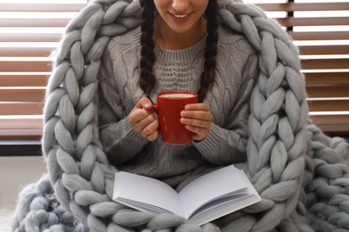 Image of Young woman wrapped in knitted blanket with cup of hot drink and book near window. Lazy morning