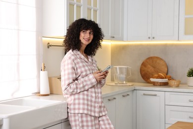 Photo of Beautiful young woman in stylish pyjama with smartphone in kitchen