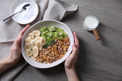 Woman holding bowl of tasty granola with banana and kiwi at grey wooden table, top view