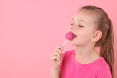 Cute little girl licking lollipop on pink background, space for text
