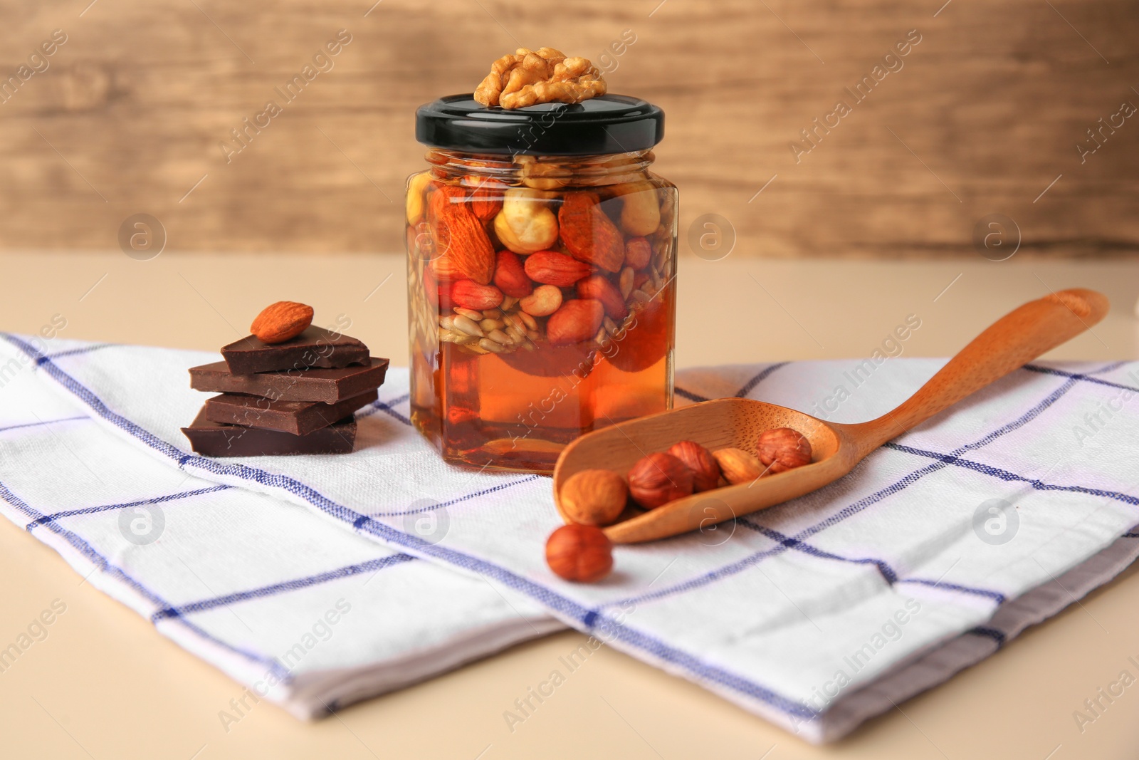 Photo of Different nuts with honey in jar, spoon and chocolate on beige table