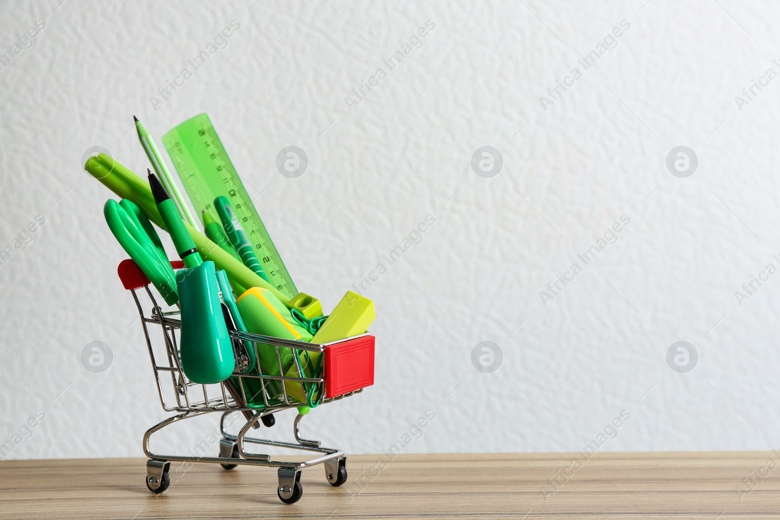 Photo of Small shopping cart with different school stationery on wooden table against white background. Space for text