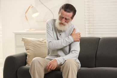 Photo of Senior man suffering from pain in arm on sofa at home. Rheumatism symptom