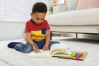 Cute African-American boy drawing in sketchbook with colorful markers on floor at home