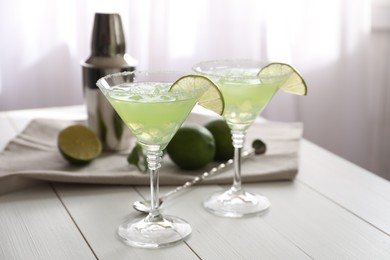 Photo of Delicious Margarita cocktail in glasses, limes, shaker and bar spoon on white wooden table