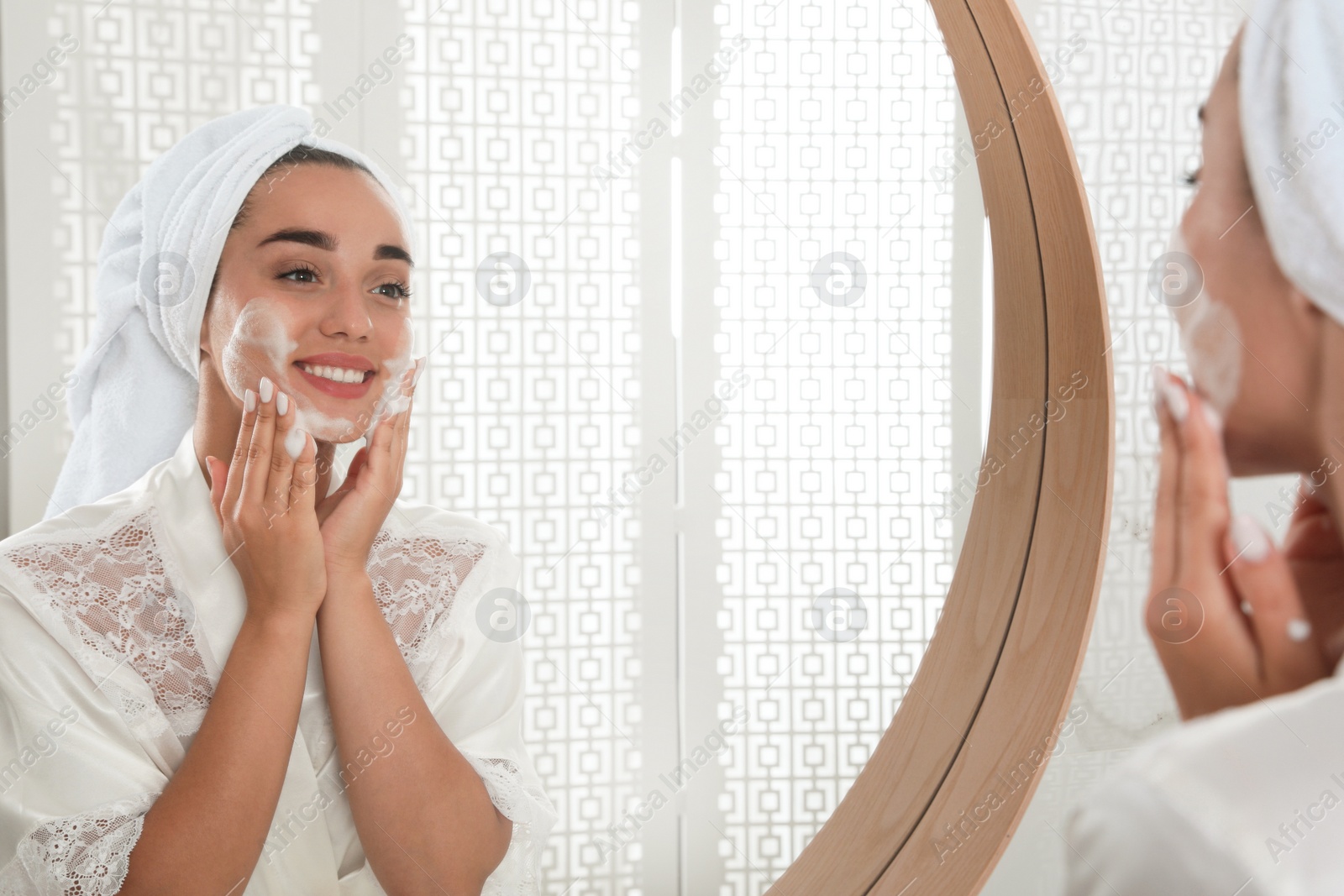 Photo of Beautiful young woman applying cleansing foam onto face near mirror in bathroom. Skin care cosmetic