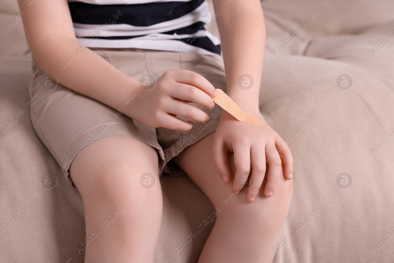 Photo of Little boy putting sticking plaster onto hand on sofa, closeup