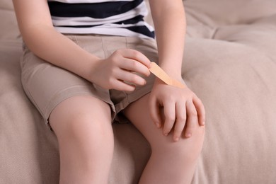 Photo of Little boy putting sticking plaster onto hand on sofa, closeup
