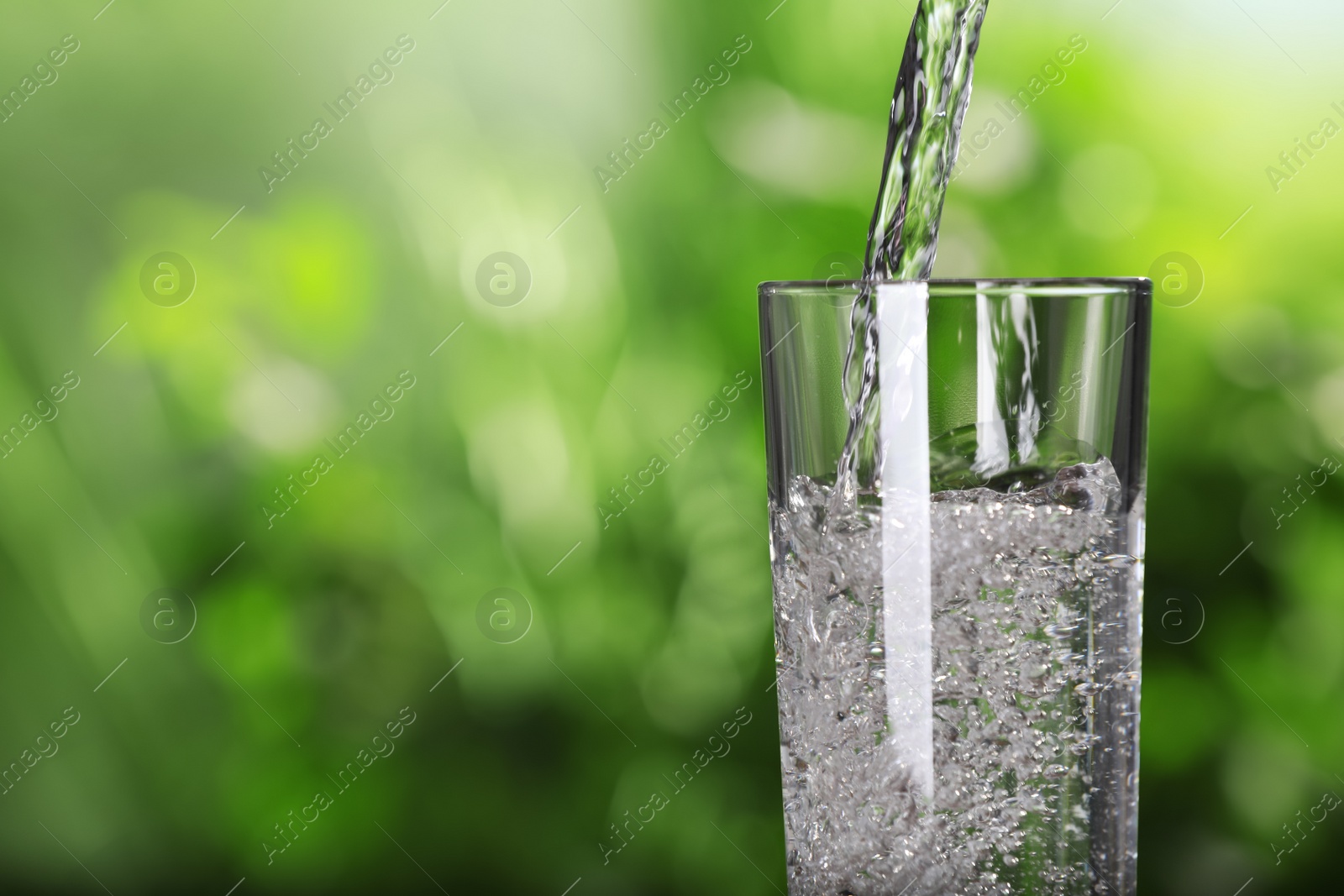 Photo of Pouring water into glass against blurred green background, closeup. Space for text