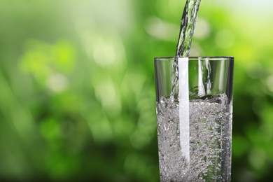 Pouring water into glass against blurred green background, closeup. Space for text