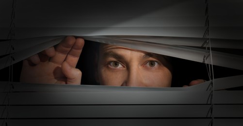 Man looking through window blinds into darkness. Paranoia concept