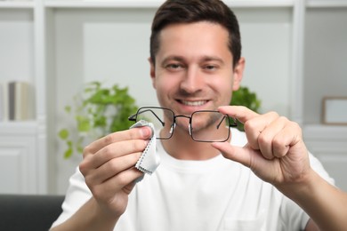 Photo of Happy man wiping glasses with microfiber cloth indoors, selective focus