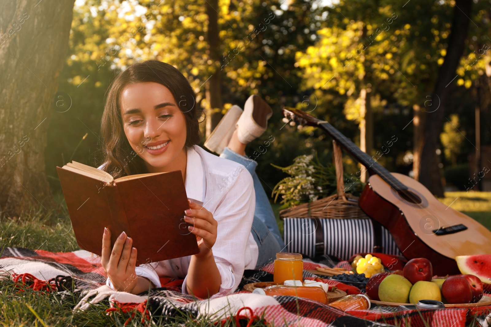 Photo of Happy young woman reading book on plaid in park. Summer picnic