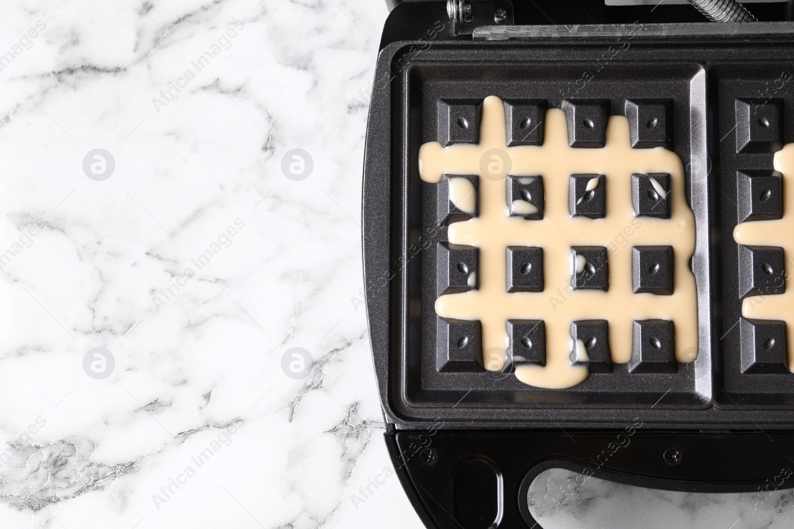 Photo of Belgian waffle maker with dough on white marble table, top view. Space for text