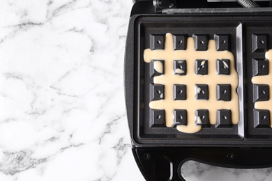 Photo of Belgian waffle maker with dough on white marble table, top view. Space for text