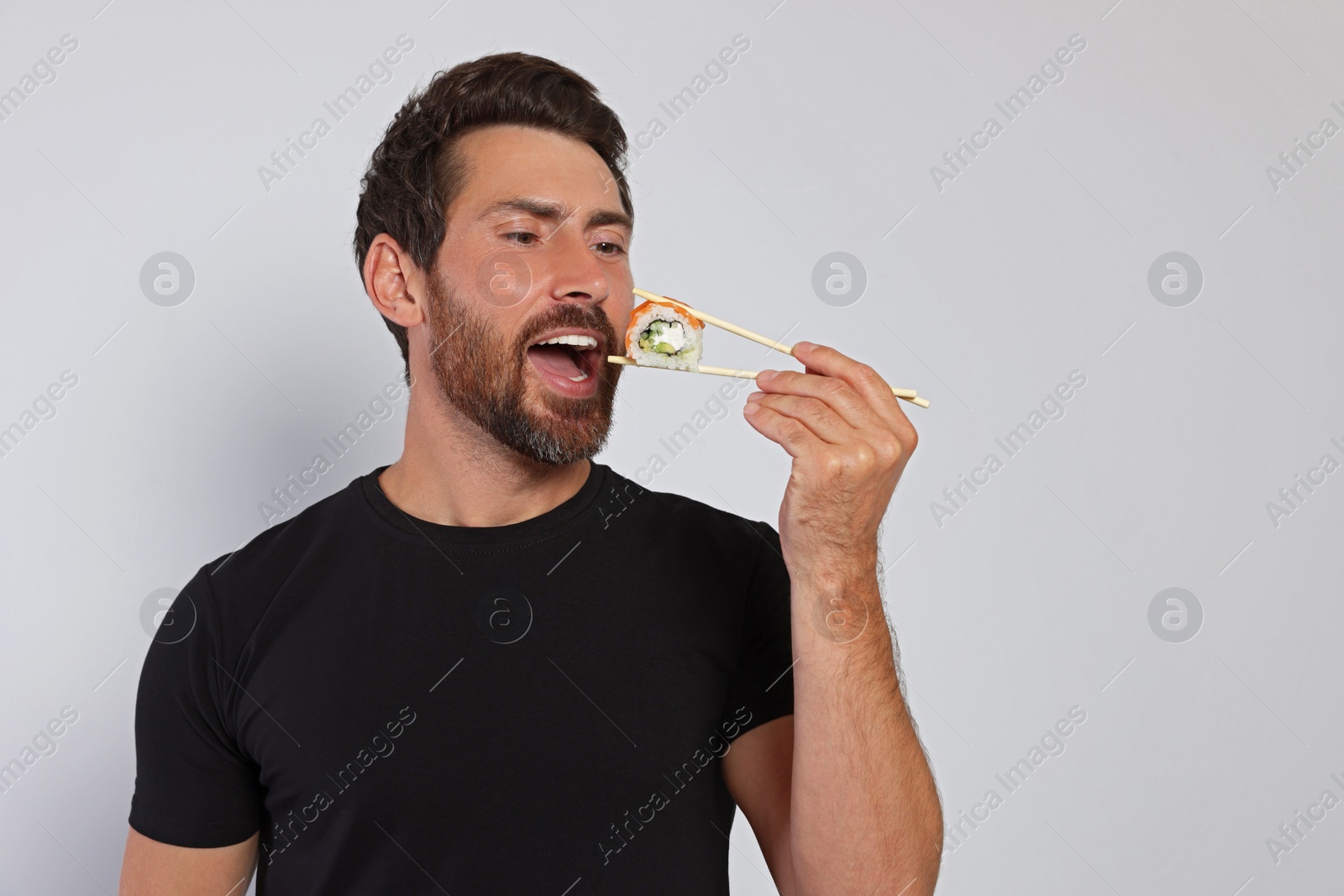 Photo of Handsome man eating sushi roll with chopsticks on light grey background. Space for text